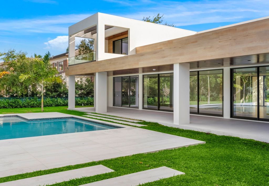 A modern two-story house with large glass windows and clean lines, crafted by a custom home builder, featuring a swimming pool, manicured lawn, and a spacious patio area under clear blue skies.