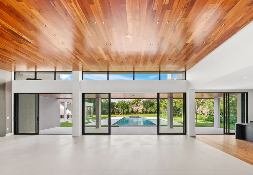 Spacious modern room designed by a custom home builder, featuring a wooden ceiling, large floor-to-ceiling glass doors, white tile flooring, and a view of the backyard pool and green garden.