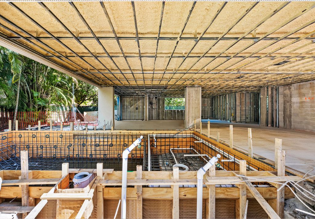 A building under construction with exposed rebar, concrete, wooden framing, and plumbing pipes. Large open rooms and a partially built pool are visible. Trees and construction materials are seen outside.