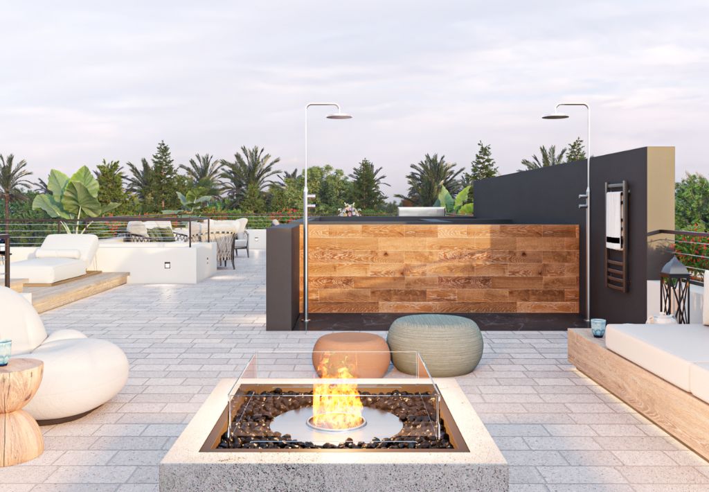 Modern rooftop patio with lounge seating, poufs, a central fire pit, wooden bar, and greenery in the background under a partly cloudy sky.