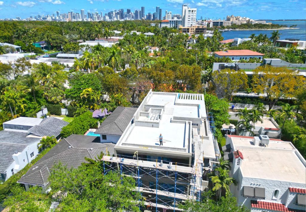 Aerial view of a house under construction with scaffolding, surrounded by trees and neighboring homes, with a city skyline and water in the background on a sunny day.