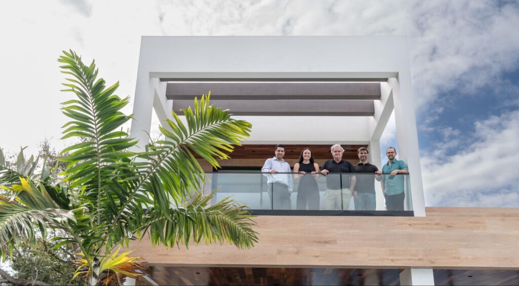 Five people stand on the balcony of a modern building designed by a Florida Keys architect, featuring white geometric lines, wooden accents, and a glass railing. A large palm plant and partly cloudy sky complete the scene.