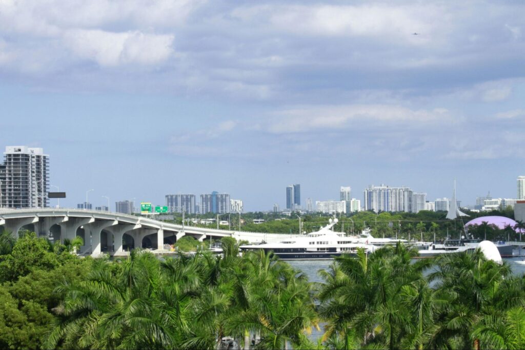A cityscape with palm trees in the foreground, a bridge crossing a waterway, and a large white yacht—showcasing high-rise buildings designed by a Florida Keys architect beneath a partly cloudy sky.
