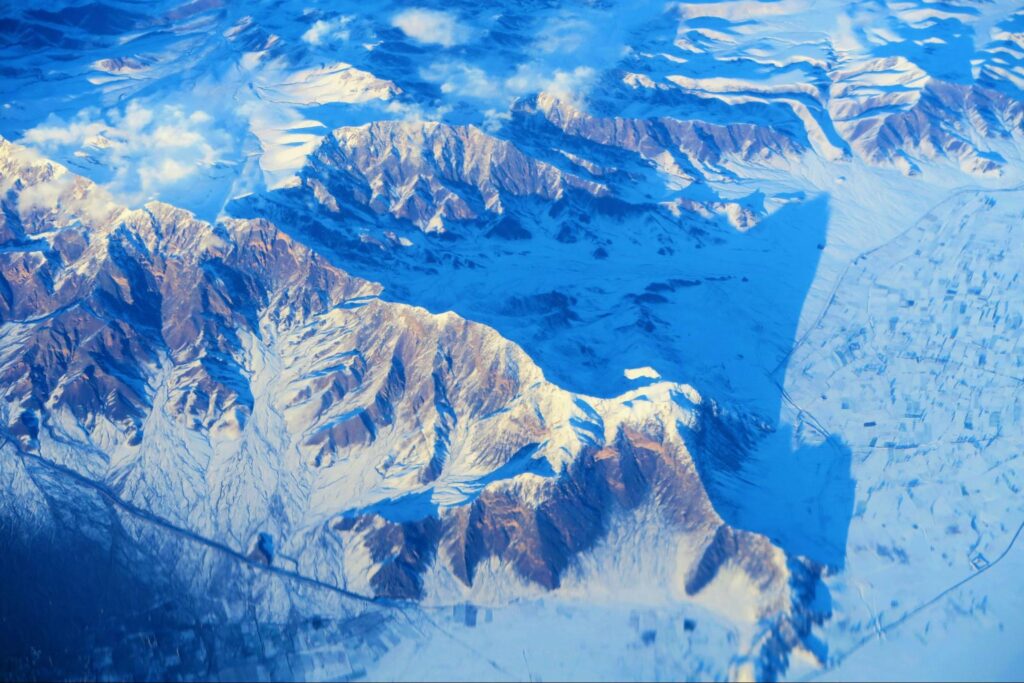 Aerial view of snow-covered mountains with rugged peaks and valleys, partly bathed in sunlight and shadow, under a clear blue sky, reminiscent of a florida keys architect blending natural beauty with striking contrasts.