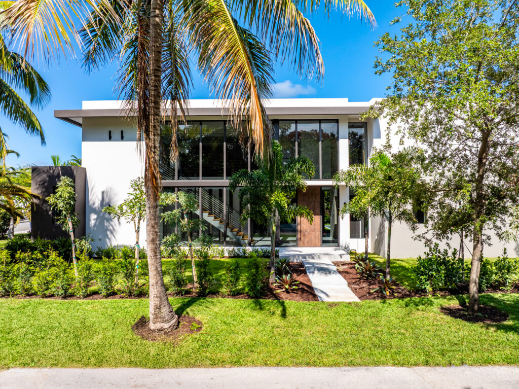 A modern two-story house in South Miami with large glass windows, surrounded by palm trees and lush green landscaping, under a clear blue sky.