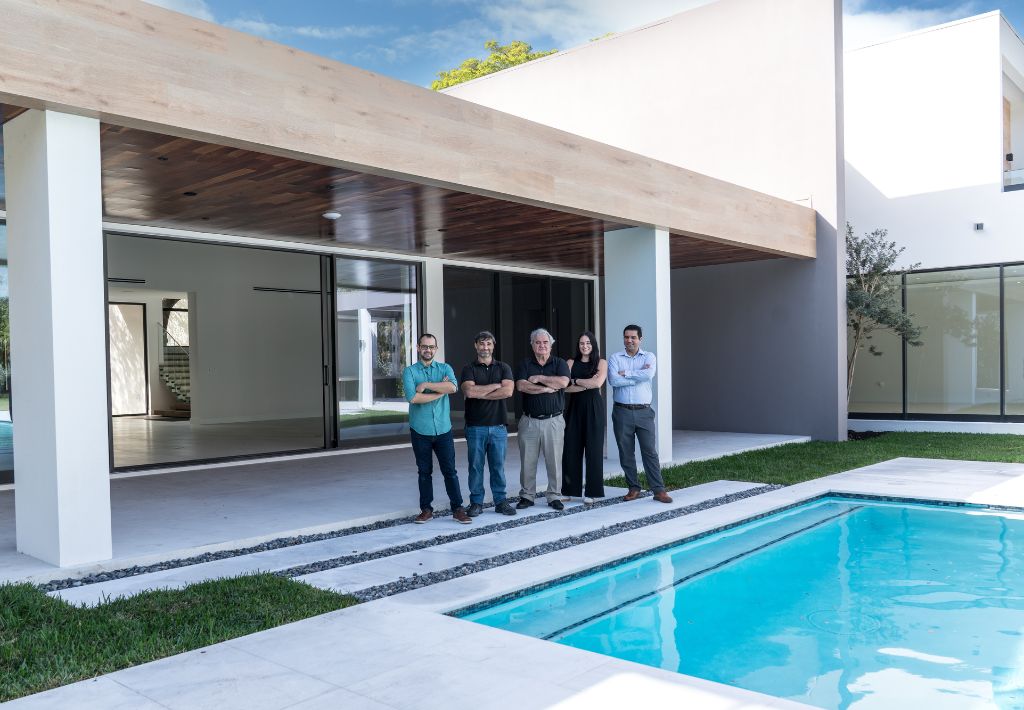 Five people stand side by side with arms crossed outside a modern house, next to a swimming pool—showcasing the work of a custom home builder—with large glass doors and minimalist architecture in the background.