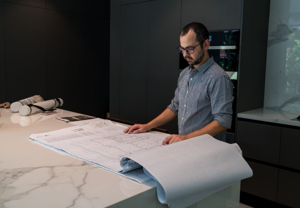 A man wearing glasses studies large architectural blueprints on a marble kitchen island, reflecting the precision of a custom home builder, with dark cabinets and rolled-up plans in the background.