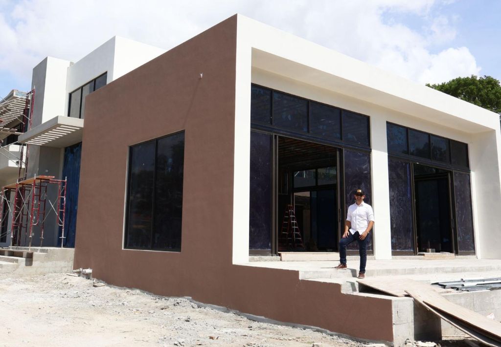 An architect stands outside a modern building designed by an ROI-focused architect, featuring large glass windows and brown and white walls. Construction materials and scaffolding surround the partially completed structure under a partly cloudy sky.