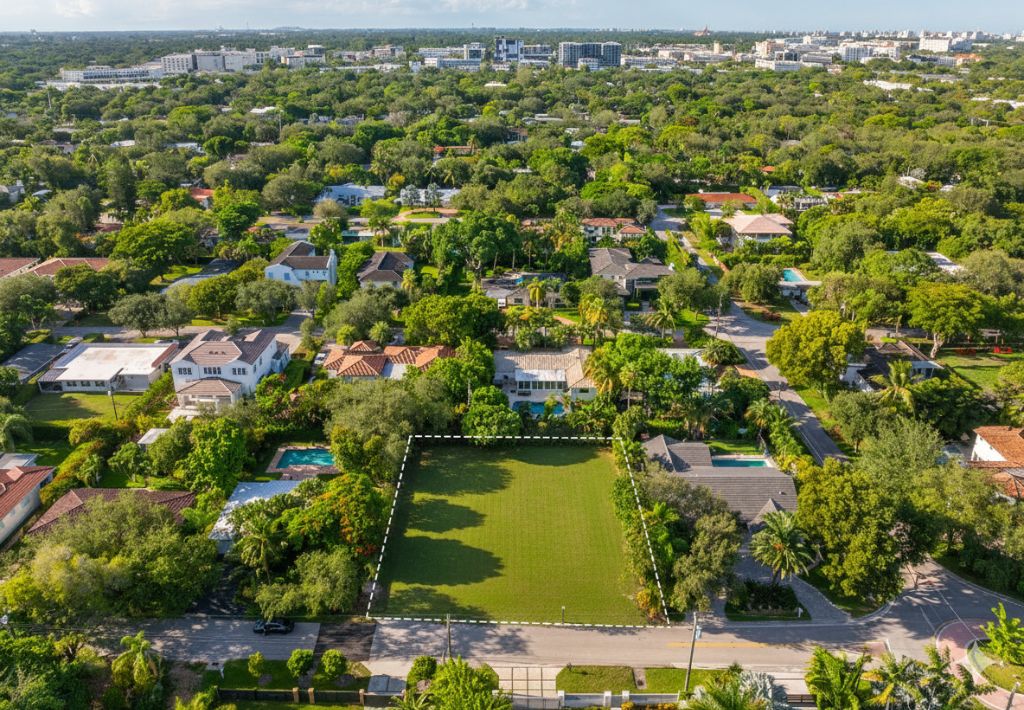 Aerial view of a residential neighborhood with tree-lined streets. In the center, a rectangular, empty green lot is outlined, surrounded by houses with pools and lush landscaping. Urban buildings are visible in the distant background and dmonstrates how to choose the right lot in Miami.