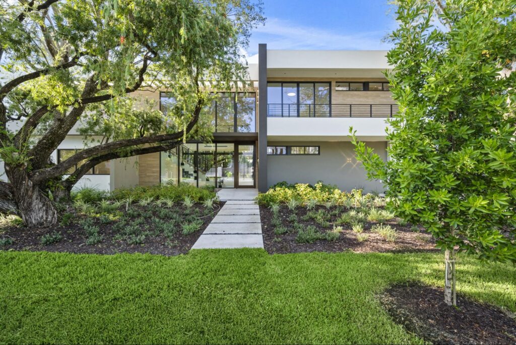 Modern two-story house with large windows, flat roof, and a sleek design, surrounded by lush landscaping, green lawn, and mature trees under a clear blue sky. This type of finished project only happens if you know how to choose the right lot to build on.