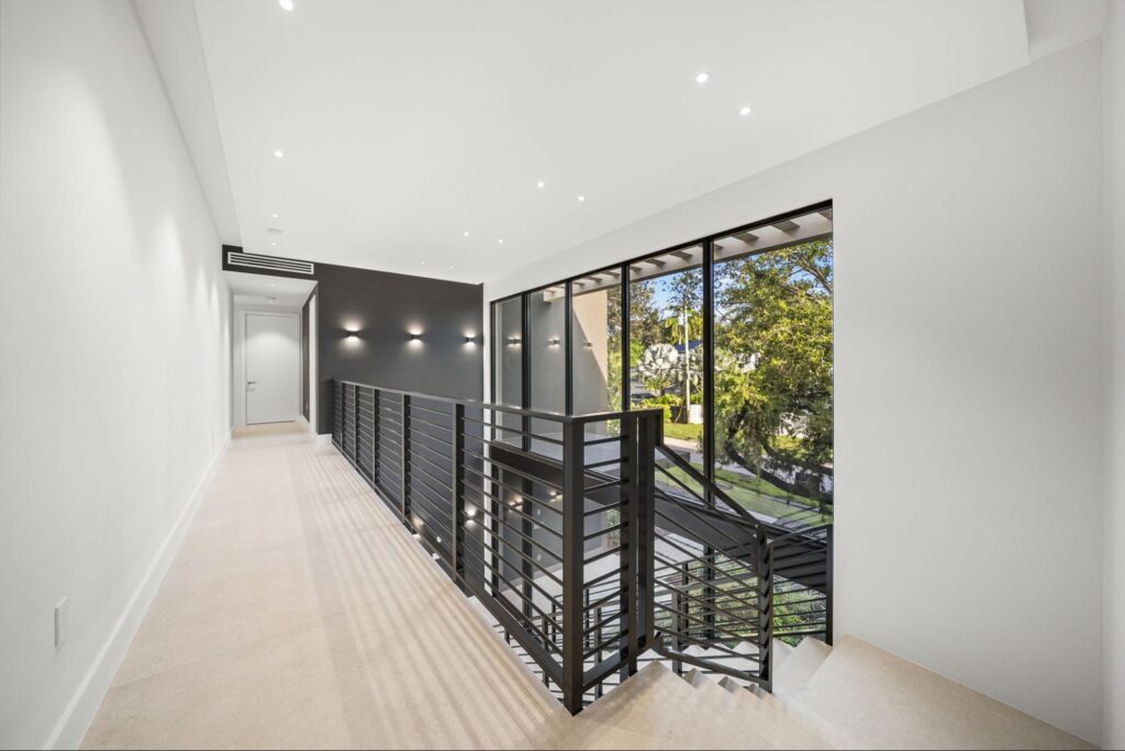 A modern indoor hallway with beige floors, white walls, black metal railing, and large windows overlooking greenery. The space is well-lit with recessed ceiling lights and an accent dark wall near the end.