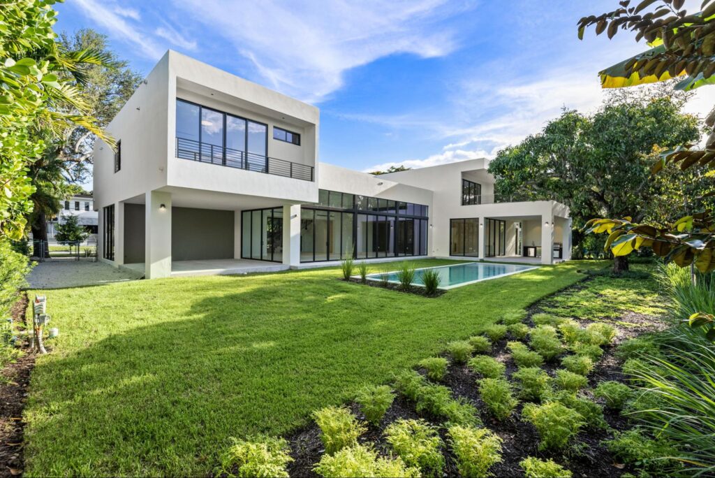 A modern two-story house with large glass windows, a flat roof, and white exterior walls sits next to a rectangular swimming pool, surrounded by a green lawn and landscaped garden. The sky is blue with scattered clouds.