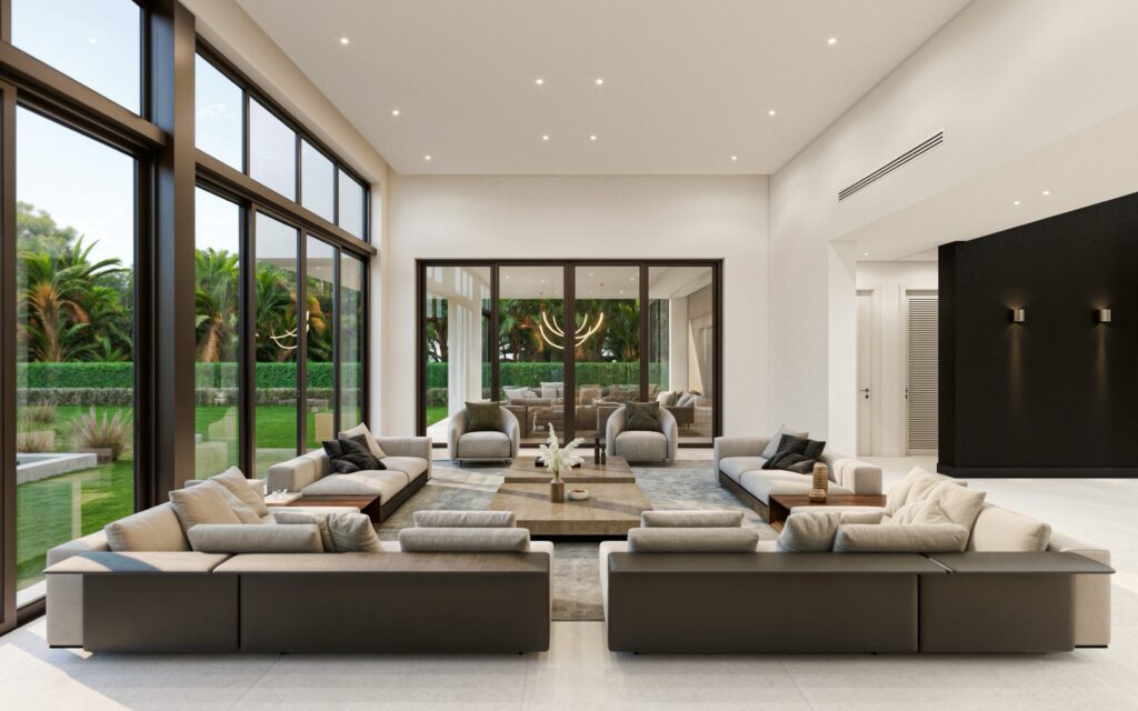 Spacious modern living room with floor-to-ceiling windows, neutral-toned sofas arranged around a coffee table, and a view of a green backyard with palm trees. A second seating area is visible through glass doors.