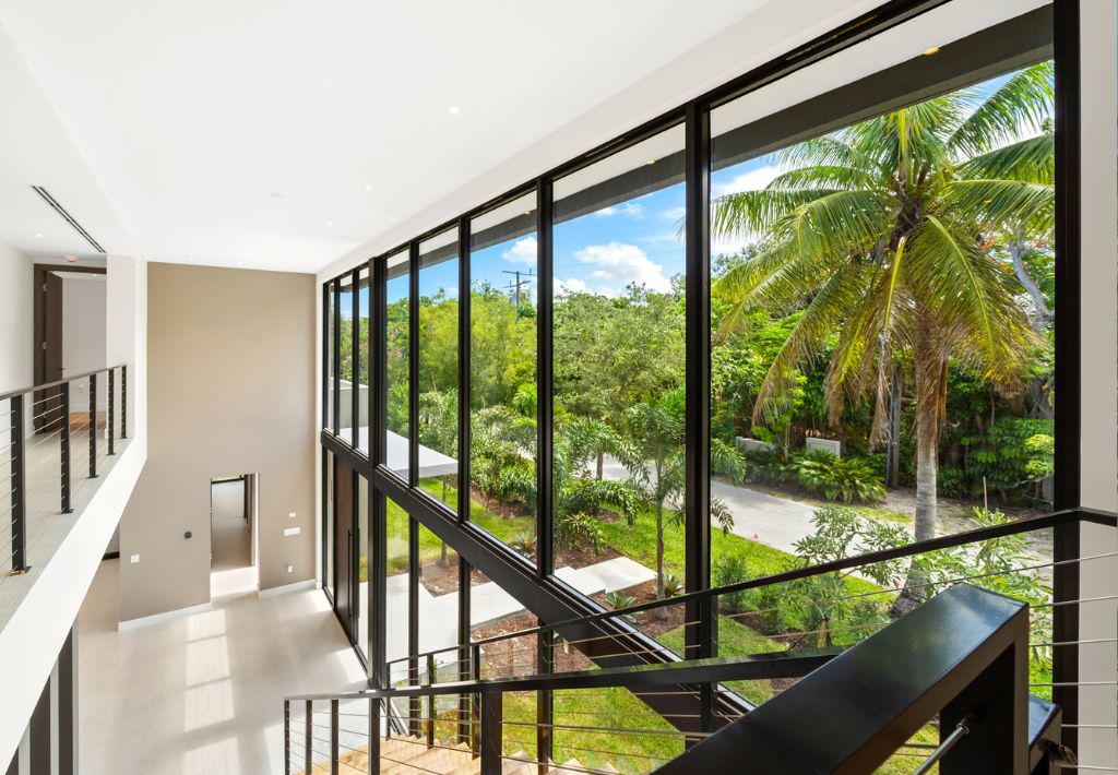 Modern interior with large floor-to-ceiling windows overlooking lush greenery and a palm tree. Black railing stairs lead down, filling the space with natural light and offering scenic resort style living at its finest.