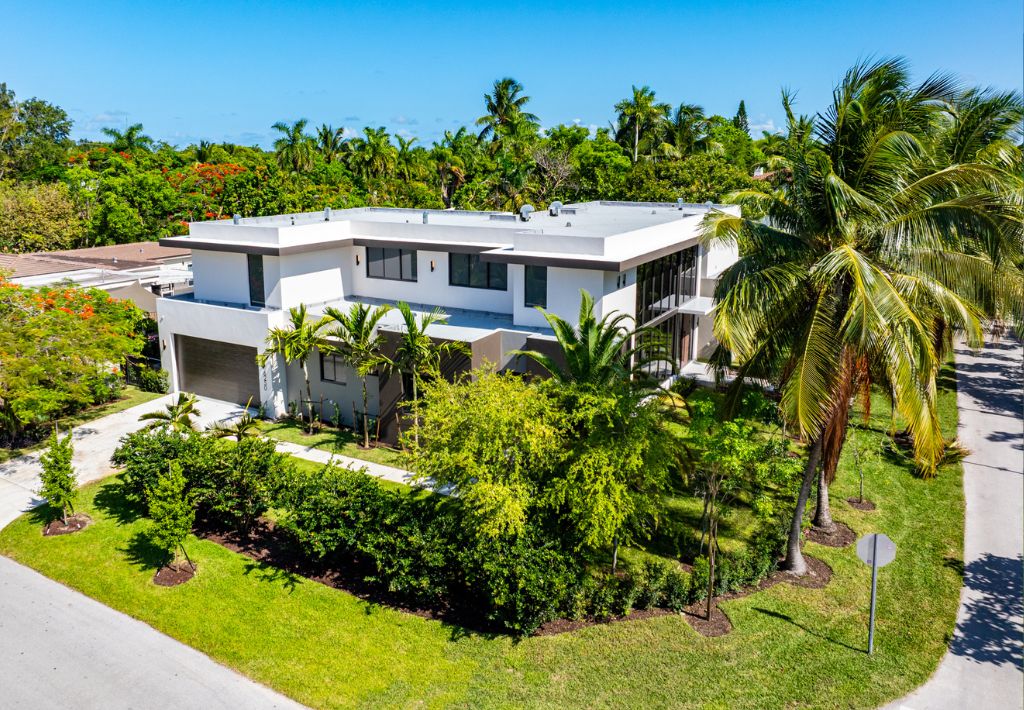 A modern two-story white house with large windows, surrounded by lush palm trees and greenery, offers resort style living on a corner lot under clear blue skies.