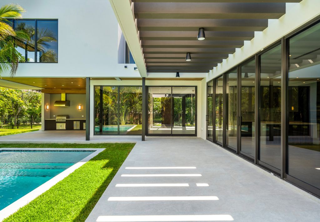 Modern house with large glass windows, covered patio, outdoor kitchen, and a swimming pool bordered by green grass—offering resort style living surrounded by trees on a sunny day.