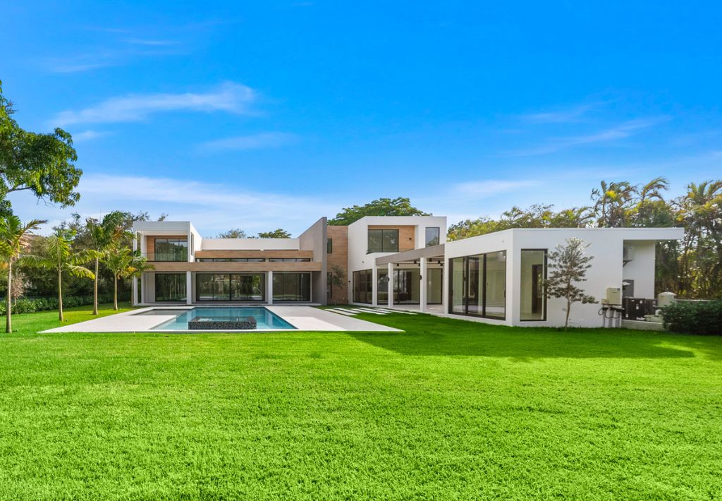 Modern two-story house with large glass windows, flat roofs, and a beige-white exterior offers resort style living, surrounded by a spacious green lawn. A rectangular swimming pool sits in the backyard under a clear blue sky.