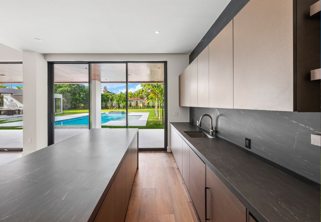 Modern kitchen with matte cabinets, dark countertops, and a large island. Floor-to-ceiling windows offer a view of the backyard pool and palm trees, bringing in natural light and creating a bright, resort style living atmosphere.