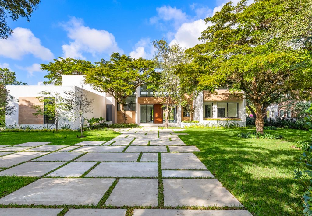 A modern two-story house with large windows, surrounded by lush green trees and grass. Enjoy resort style living with a geometric stone pathway in the front yard, leading to the entrance under a bright blue sky with scattered clouds.