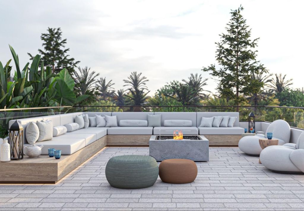 Modern outdoor lounge area with light gray sectional sofas, round poufs, and a central fire pit on a tiled patio, surrounded by lush greenery and tall trees—perfect for resort style living.