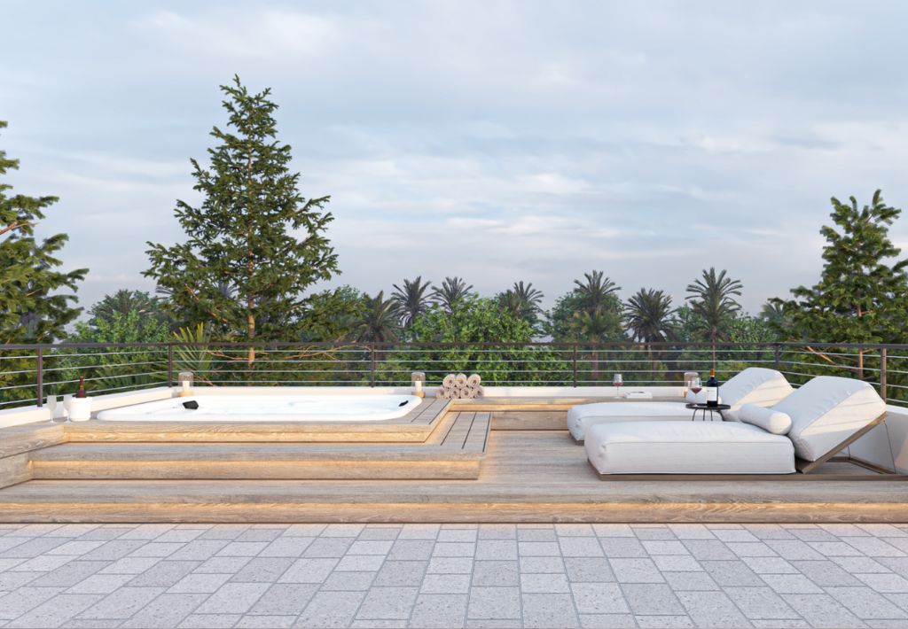 A modern outdoor patio offers resort style living with two lounge chairs, a small table, and a built-in hot tub, all surrounded by greenery and trees under a cloudy sky. Towels and drinks are set beside the hot tub and chairs.