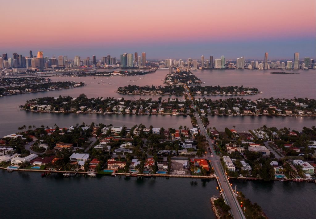 Aerial view the Venetian Islands, an investment alternative to star island in Miami. The city skyline is in the distance under a pink and blue sunset sky, likely Miami Beach and downtown Miami.