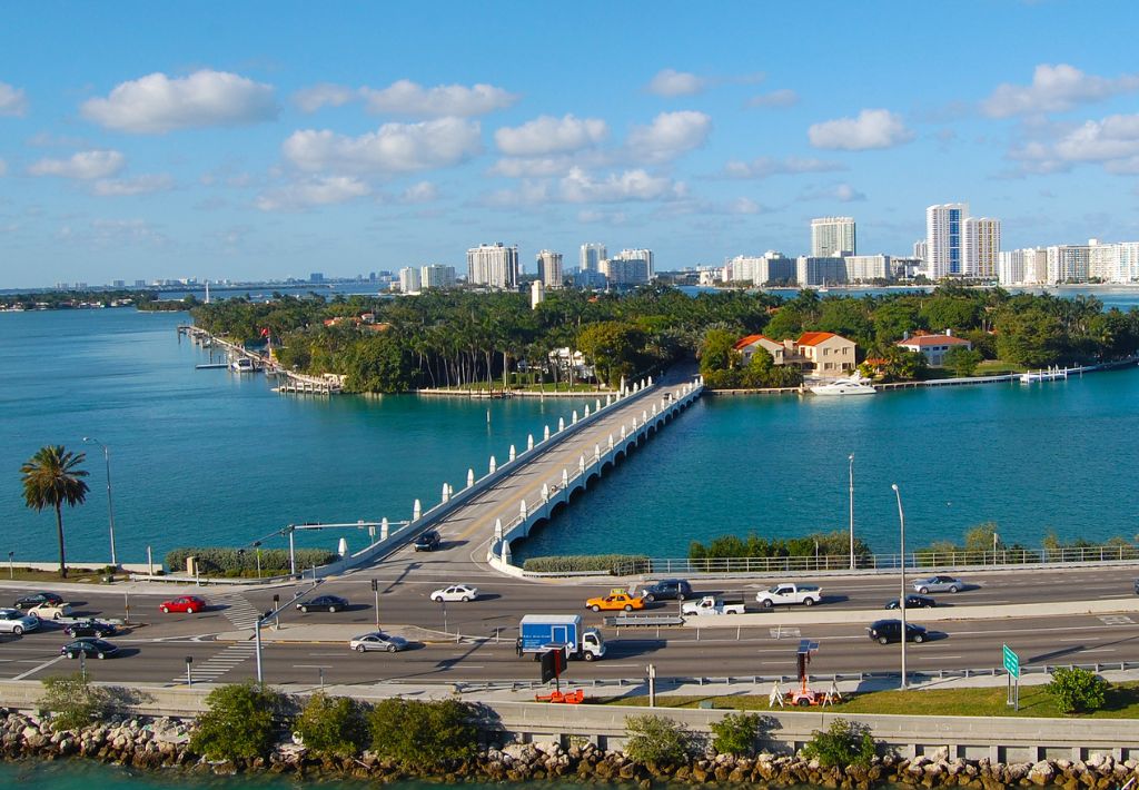 A scenic view of a causeway lined with palm trees connecting the mainland to Star Island in Miami, with waterfront homes, surrounded by blue water and high-rise buildings in the background under a clear sky.