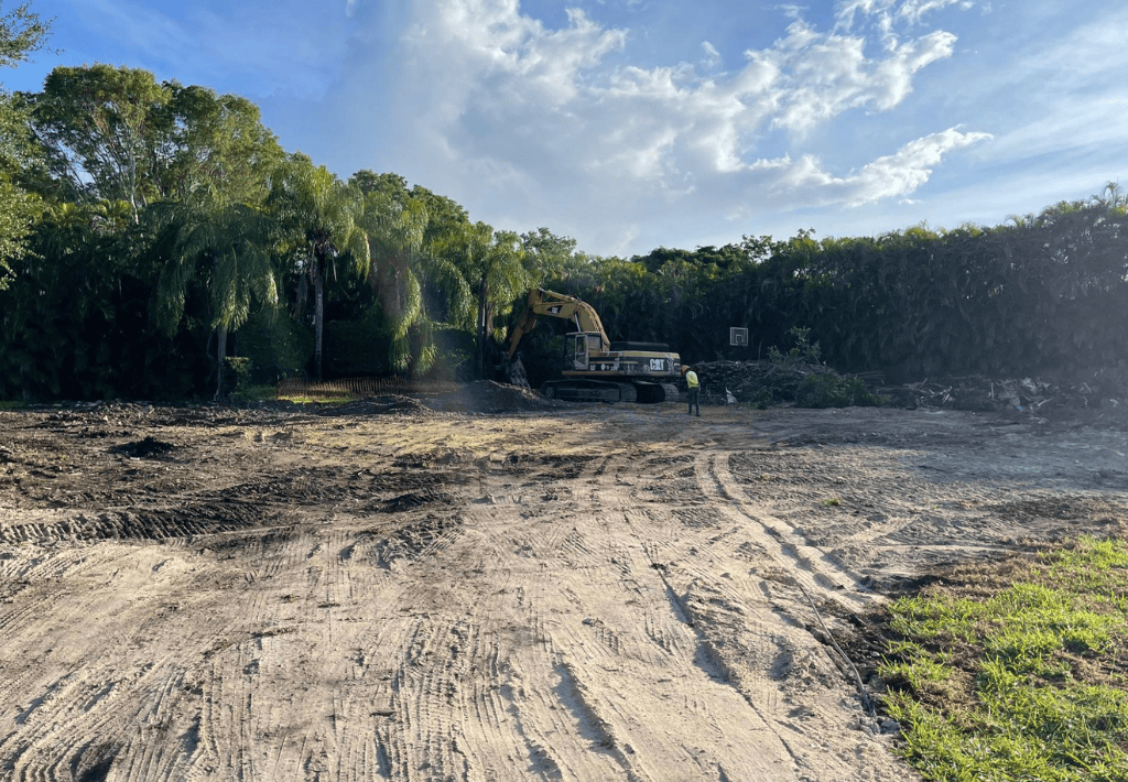 A construction site with a yellow excavator and a worker wearing a helmet, surrounded by trees and a mostly clear sky with some clouds overhead. The ground is dirt with tire and track marks. This is the first phase of ground up new construction.
