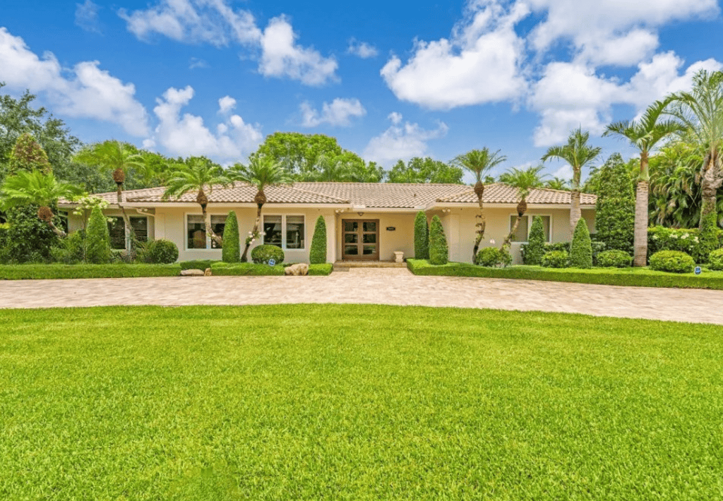 Single-story house with large windows, tan roof, and manicured landscaping, surrounded by palm trees and lush green lawn under a bright blue sky with scattered clouds. This is what the property looked like prior to ground up new construction.