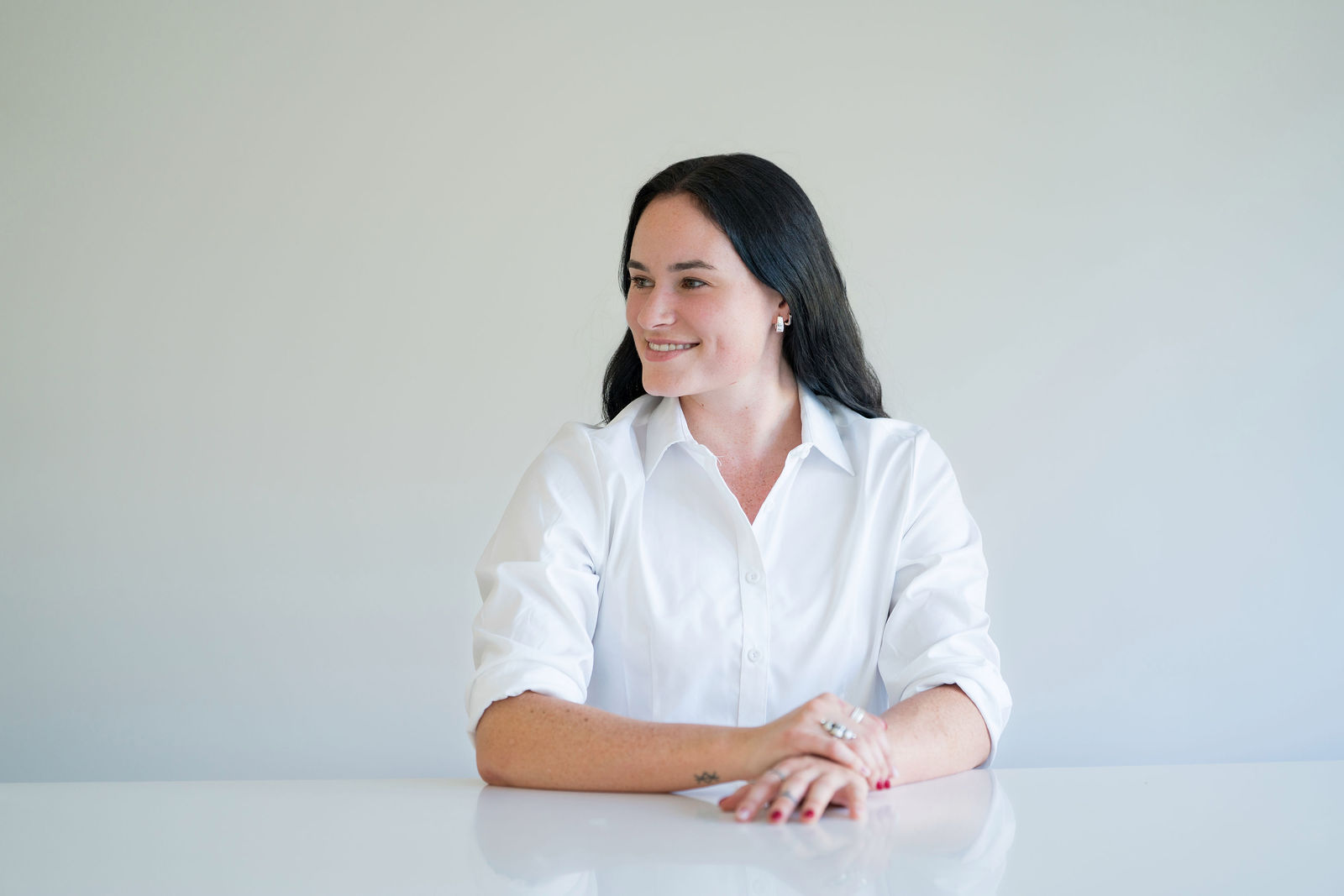 A woman with long dark hair wearing a white button-up shirt sits at a white table, smiling and looking to her right against a plain light background.