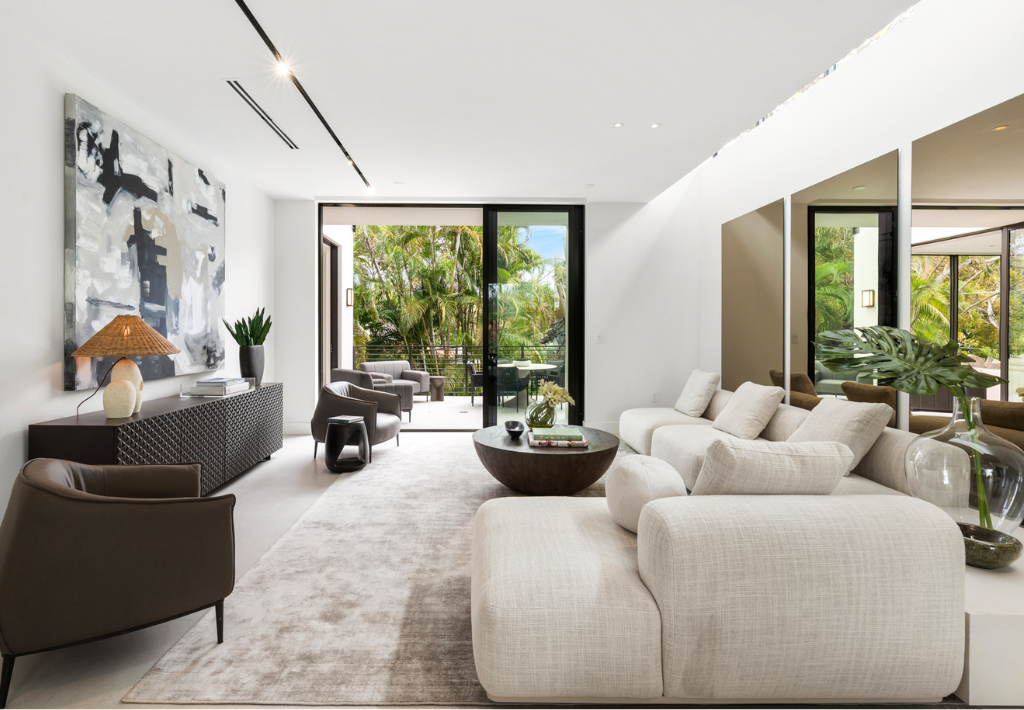 An Interior Designer in Miami has furnished A modern living room with white sofas, a round coffee table, neutral decor, large abstract art, and glass doors opening to a lush green outdoor space with natural light filling the room.