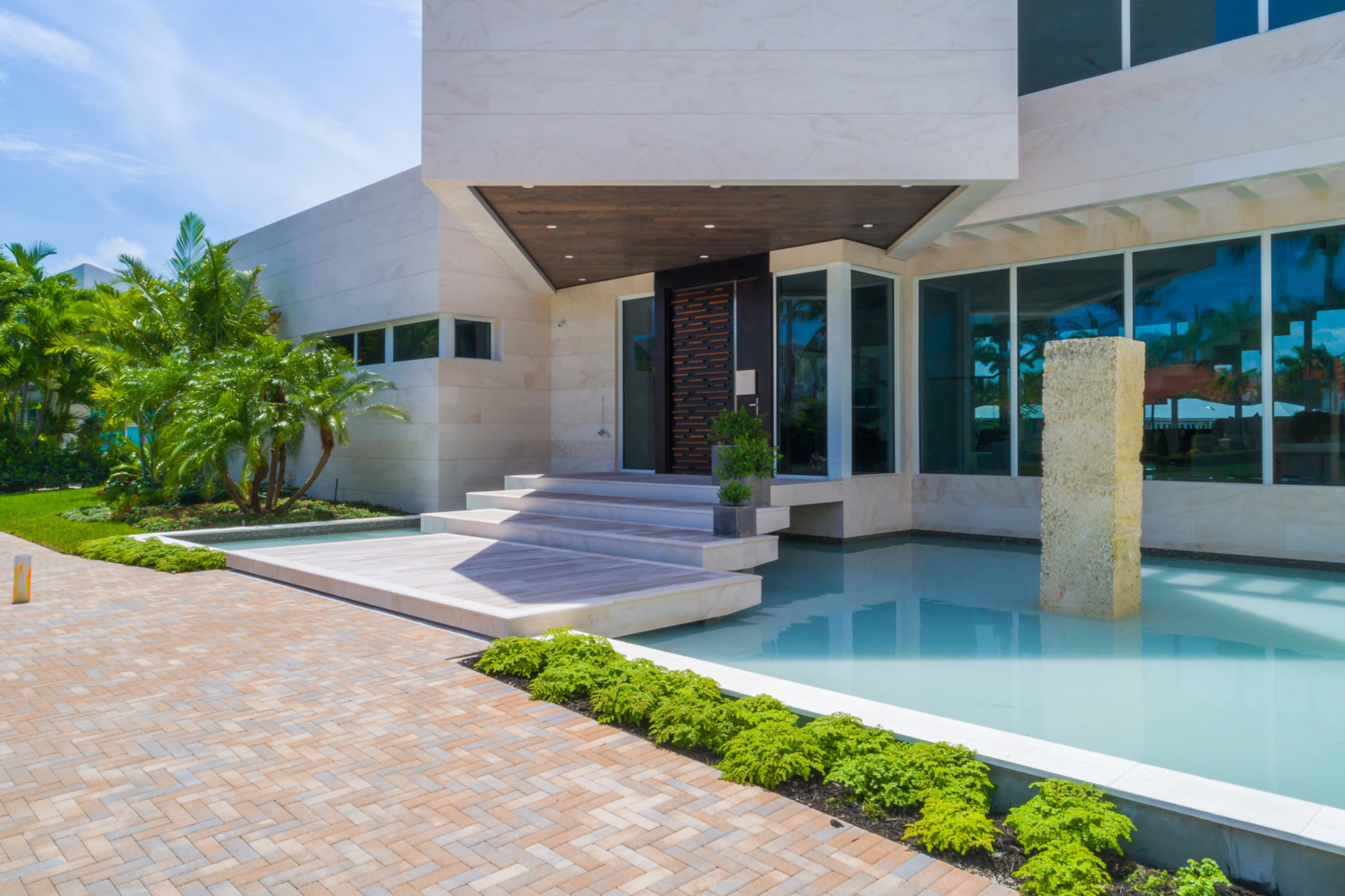 Modern house entrance with large steps, surrounded by water feature and green landscaping, featuring a unique front door and large glass windows reflecting palm trees and blue sky. This rotated feature is iconic in current coral gables architecture examples.