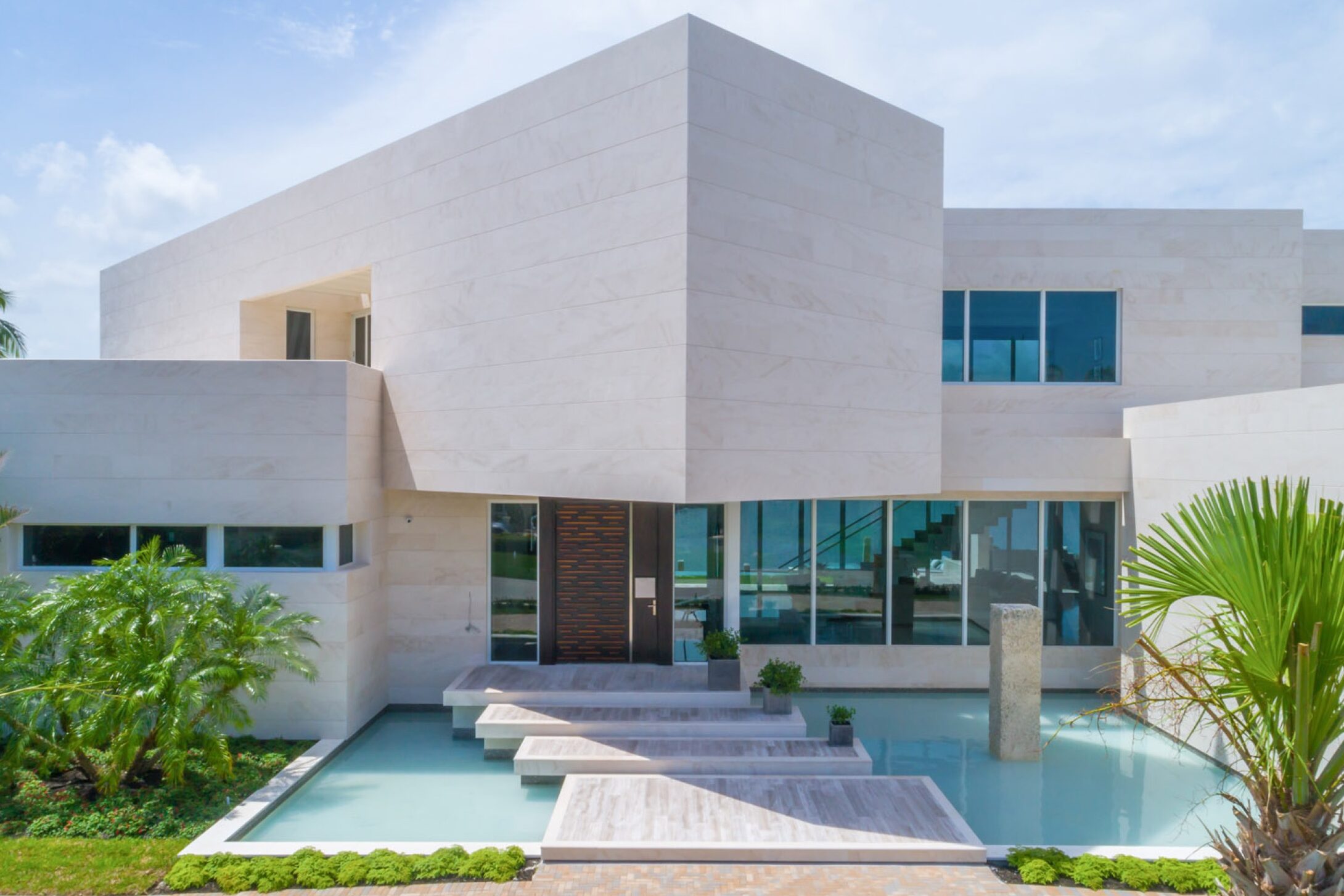 Modern white stone house with geometric architecture, large windows, and a wooden front door. A floating walkway crosses a shallow reflecting pool, surrounded by tropical plants and palm trees under a partly cloudy sky. This is one of the more distinctive coral gables architecture examples with stone from a spanish quarry paying tribute to the spanish style homes dominating the neighborhood.