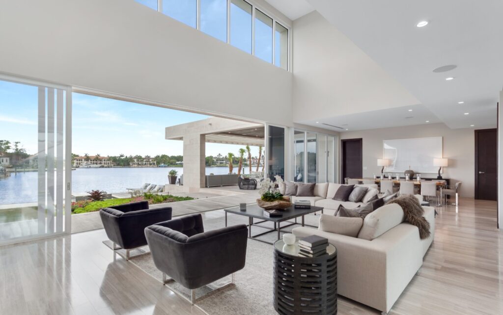 Modern living room with floor-to-ceiling windows, neutral-colored furniture, and an open layout. Designed by an interior designer, the space opens to a patio with lake views and lush greenery, creating a bright and airy atmosphere.