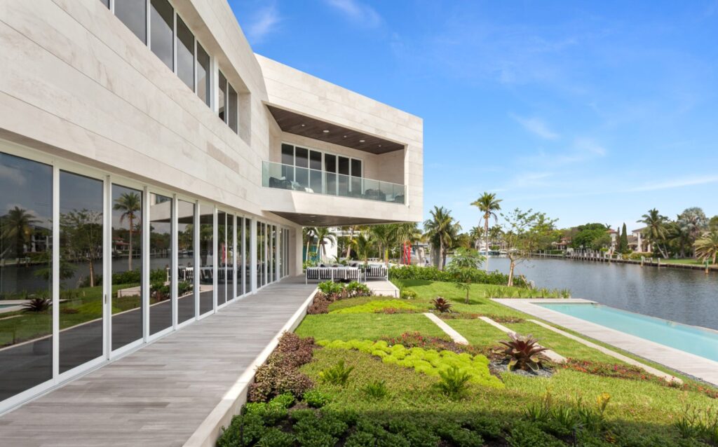 A modern house with large glass windows, a balcony, and a spacious patio overlooks a lush green garden and a canal lined with palm trees under a clear blue sky with other Coral Gables Architecture styles seen across the intercoastal.