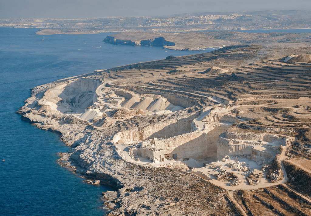 Aerial view of a coastal limestone quarry with terraced excavation levels near the blue sea, surrounded by rugged terrain and distant landforms under a clear sky. Solano Prado is an example of coral gables architecture that utilizes materials right from the premium source.
