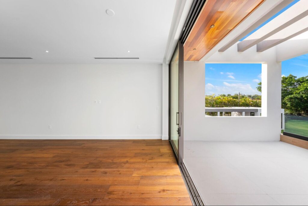 Modern, empty room with wooden flooring and white walls opens to a spacious balcony with large sliding glass doors. The balcony has a wooden ceiling, white floor, and scenic outdoor views of trees and blue sky. There is no furniture here but an Interior Designer with a flair for modern design will know just what to do.