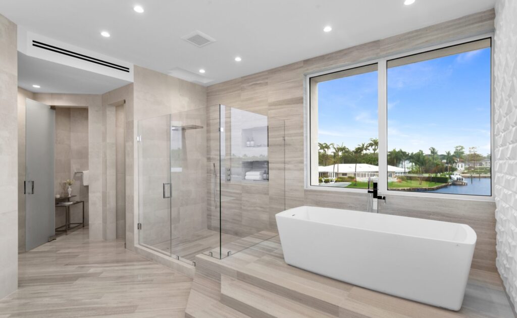 Coral Gables Architecture example featuring a Modern bathroom with a freestanding white bathtub by a large window, a glass-enclosed shower, beige tile flooring and walls, and a view of palm trees and water outside.