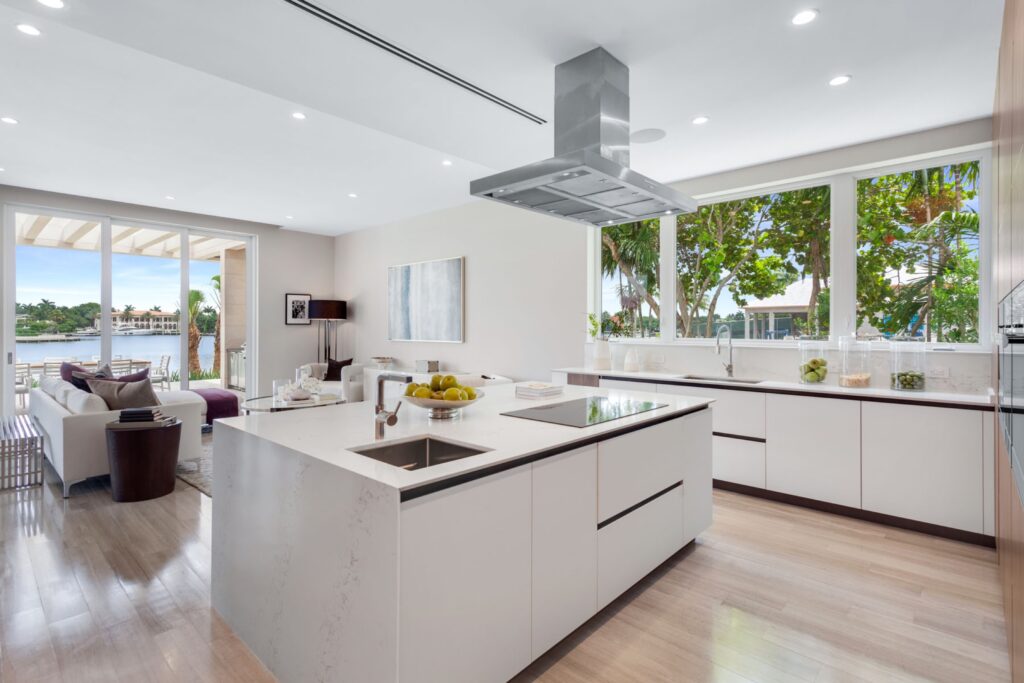 Modern open-concept kitchen with a large white island, stainless steel appliances, sink, and stove. Large windows provide a view of greenery and water outside. The living area is visible in the background. Distinctive Coral Gables Architecture all the way.