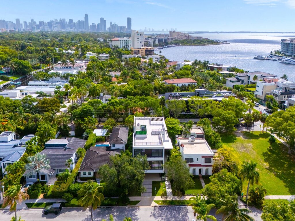 Aerial view of a coastal city neighborhood with luxury homes in Miami, lush green trees, waterfront properties, and a city skyline visible in the distance under a clear blue sky.