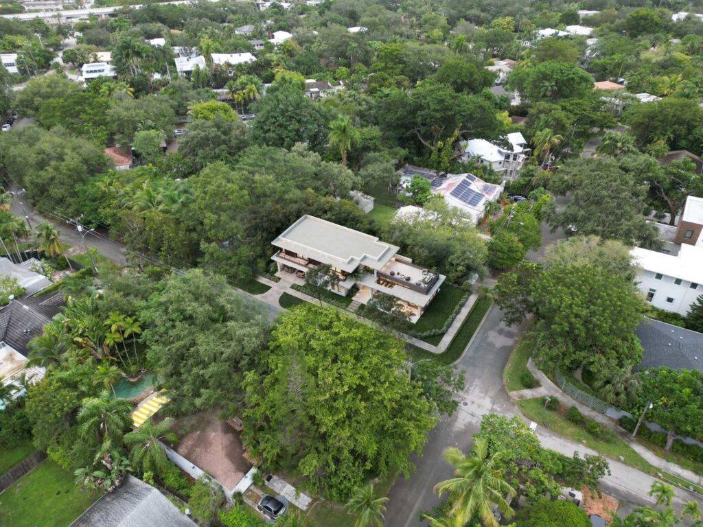 Aerial view of a modern, flat-roofed house surrounded by lush trees, located at the intersection of two residential streets in a green suburban neighborhood, exemplifying the elegance found in luxury homes in Miami.