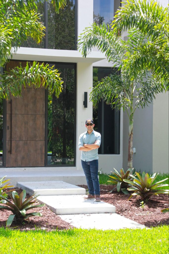 A man wearing sunglasses, a cap, and casual clothes stands with his arms crossed on a pathway in front of a modern house surrounded by tropical plants and greenery, capturing the relaxed elegance often seen at luxury homes in Miami.