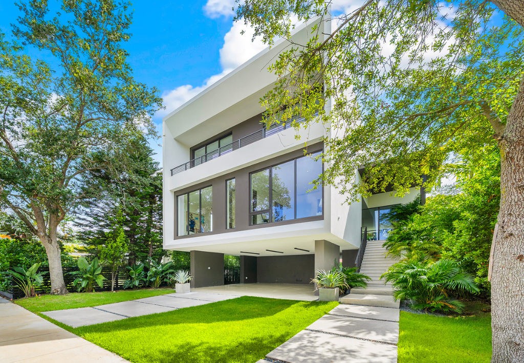 A modern, white, two-story house with large windows, surrounded by lush greenery and trees, featuring an elevated structure and a neatly manicured lawn under a bright blue sky showing how warm modern homes can truly be.