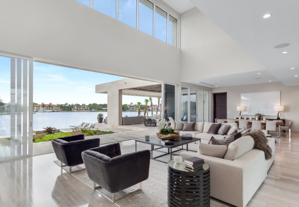 Modern living room with large windows, neutral furniture, and open layout, overlooking a patio and waterfront view with palm trees and buildings across the water. Dining area visible in the background where modern homes like this demonstrate luxurious living.