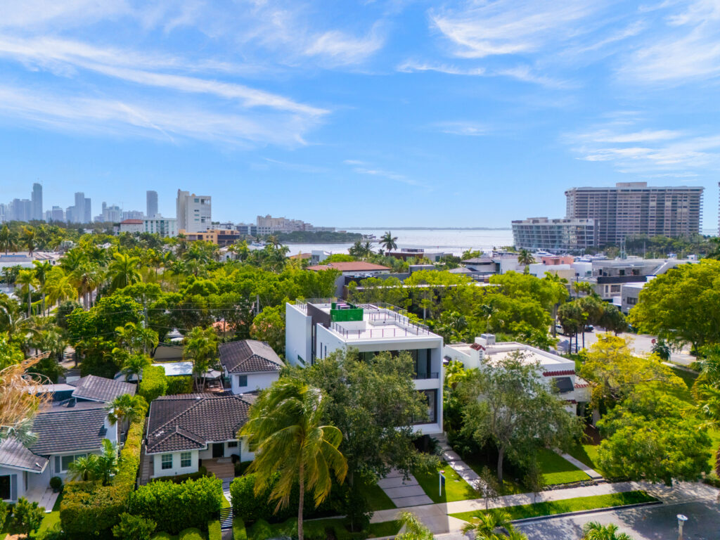 Aerial view of a residential neighborhood with modern white homes, lush green trees, and a coastal city skyline in the distance illustrating the diverse settings available for resudential life in miami.
